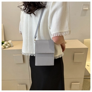 Close-up of a minimalist grey glossy leather crossbody bag worn on a white fringe top