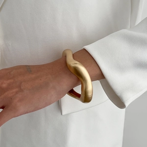 Detail view of a matte gold wavy wide bracelet paired with a stylish white blazer sleeve
