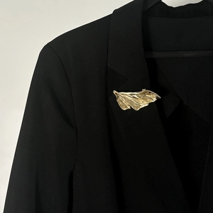 Dettaglio spilla a piuma dorata con trama metallica pieghettata su un blazer nero