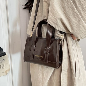 Dark brown glossy small square shoulder bag with gold lettering shown on light trench coat