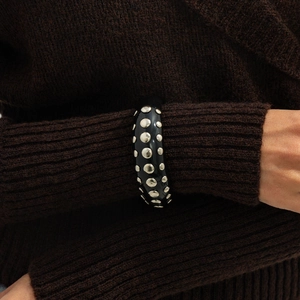 Detail of a wide black resin cuff with silver rivets on a brown knit sweater sleeve