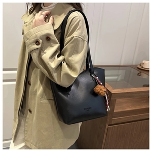 Detail of black pebbled tote bag with plush bear charm on a khaki trench coat