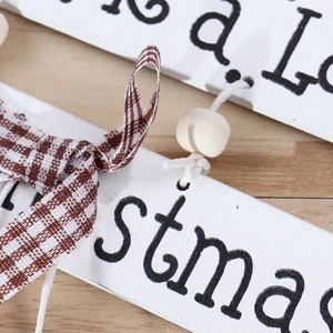 White wooden Christmas tree ornament with black lettering, plaid ribbon, and wood beads