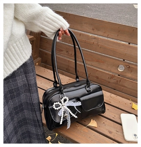 Glossy black tote bag with flap pockets and pearl bow charm shown on a park bench