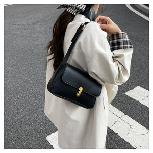 Detail of a black leather shoulder bag with a gold oval clasp worn by a model outdoors