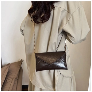 Detail of a dark brown glossy envelope crossbody bag with a gold chain on a model
