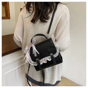 Detail of a black square crossbody bag with white lace trim and bow on a beige sweater