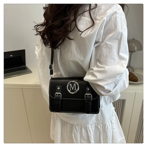 Close-up of black crossbody bag with silver M logo worn by model in white dress