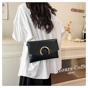 Modern black envelope bag with gold buckle shown on shoulder in a bright cafe setting