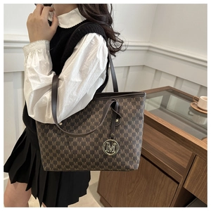 Brown tote bag with M-pattern print and gold pendant worn by model in a cozy cafe
