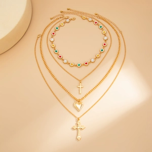 Dettaglio collana a strati dorata con ciondoli a croce di strass e cuore su sfondo beige