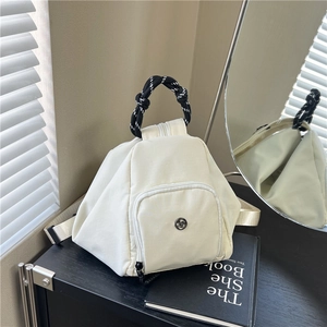 Mochila triangular crema con asa trenzada sobre un mueble negro en una habitación moderna