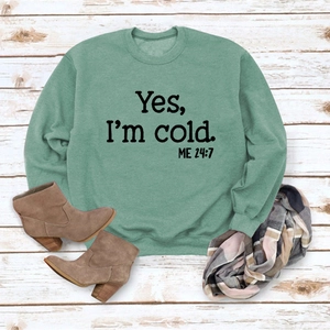 Flat lay of a green fleece sweatshirt with Yes I'm cold slogan on a wooden floor