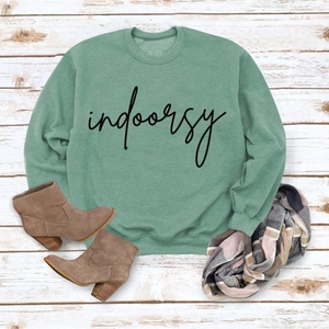 Flat lay view of a mint green sweatshirt with 'indoorsy' print on white wood background