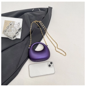 Detail of purple acrylic crescent handbag with gold chain strap beside a phone for scale