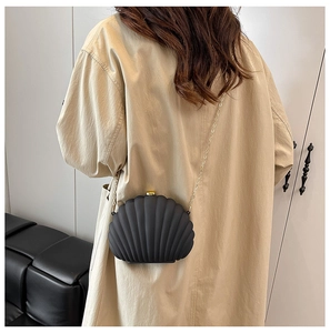 Detail of a matte black shell-shaped crossbody bag with a gold chain on a trench coat