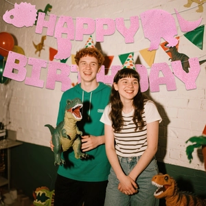 Roze glitter dinosaurus Happy Birthday banner vastgehouden door mensen op een feestje