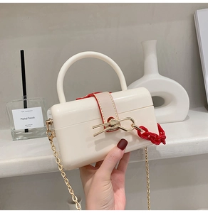 Detail of a creamy white rigid box bag featuring a red decorative chain and gold shoulder strap