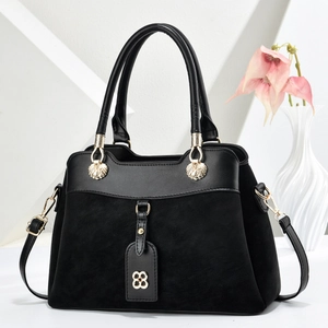 Detail of black suede handbag with gold shell hardware and floral charm on a white pedestal