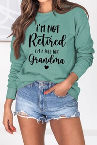 Detail of sage green sweatshirt featuring 'I'M NOT Retired' black artistic text print