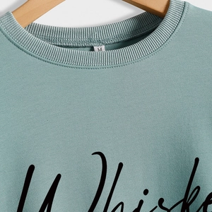 Sage green crewneck sweatshirt with Whiskey Weather letter print on a wooden hanger
