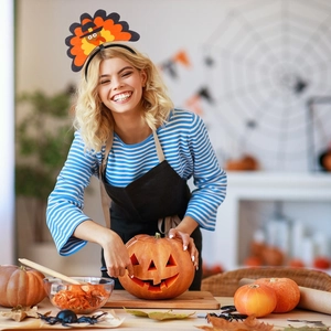 Woman wearing a festive turkey headband while carving a pumpkin in a cozy kitchen setting