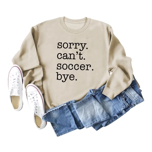 Flat lay of beige sweatshirt with Sorry Can't Soccer Bye text, jeans, and sneakers