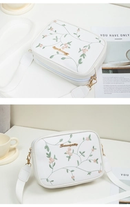 Détail de broderie florale et logo doré sur sac bandoulière blanc avec sangle large