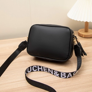 Schwarze PU-Leder Umhängetasche mit breitem Logo-Gurt und Quaste auf hellem Holztisch