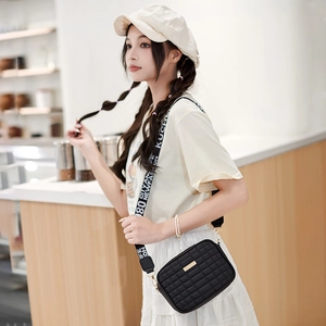 Black quilted crossbody camera bag with wide printed strap and minimalist metal plaque