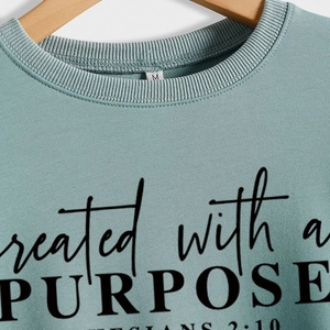 Close-up of light cyan crewneck sweatshirt with 'Created with a Purpose' print on a wooden hanger