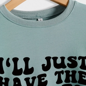 Sage green crewneck sweatshirt with 'I'LL JUST HAVE THE' text on wooden hanger