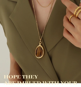 Golden teardrop tiger eye pendant necklace styled with textured ring on olive blazer