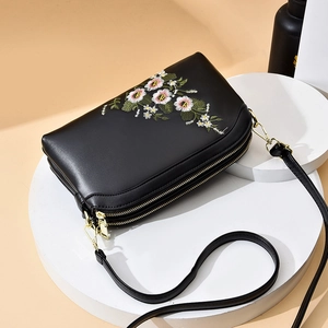 Elegant black leather crossbody bag with floral embroidery and dual gold zippers on a pedestal