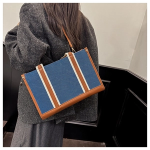Detail of a large denim tote bag with brown leather trim carried by a model in a coat
