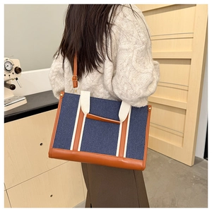 Rear view of a blue denim tote bag with brown leather trim held by a woman in a cafe