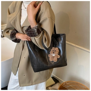 Detail of a black leather tote bag with a cute brown bear charm on a khaki trench coat