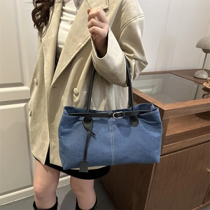 Blue denim tote bag with leather straps and decorative belt detail shown in a cozy room
