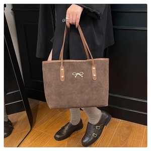 Detail of a brown suede tote bag featuring a delicate metal bow and slim leather handles