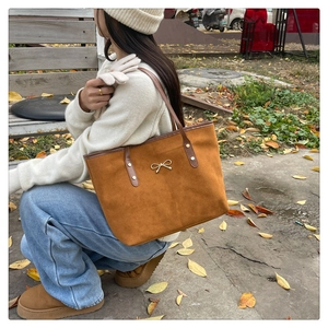Détail d'un cabas en daim marron avec nœud doré et anses en cuir dans un décor d'automne