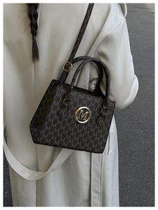 Detail of dark brown tote bag featuring M-pattern print and golden logo emblem