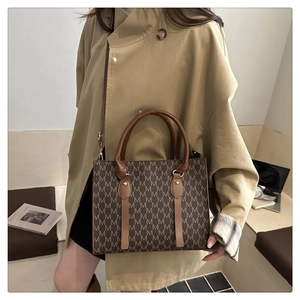 Brown tote bag with 'M' monogram and leather straps shown in a lifestyle office setting