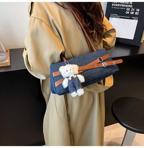 Detail of a denim shoulder bag featuring brown leather straps and a plush bear charm