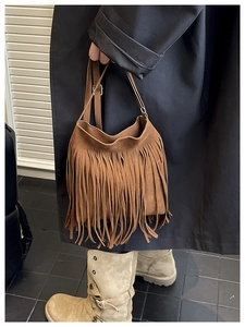 Bolso tipo cubo de ante marrón con flecos largos mostrado por una modelo con gabardina negra