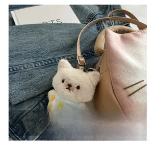 Weißer Plüschkatzen-Anhänger mit blauem Schal an einer beigen Handtasche mit Metallhaken