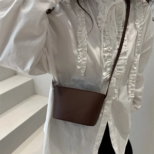 Minimalist dark coffee leather mini crossbody bucket bag shown on a white ruffled shirt
