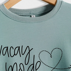 Green long sleeve sweatshirt with vacay mode text and heart airplane print on wood hanger