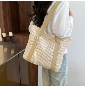 Detail of woven straw tote bag featuring white tassel charm and wide beige shoulder strap