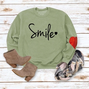 Detail of green crewneck sweatshirt with Smile print and heart accent on white wood floor