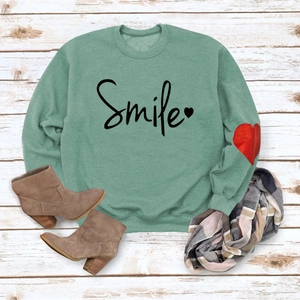 Flat lay of bean green sweatshirt featuring a Smile print and red heart elbow patch